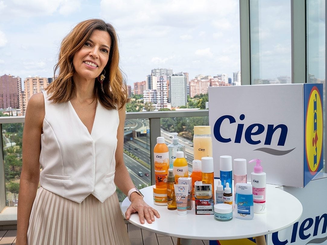 Woman in white sleeveless top standing at table with Cien skincare products and city skyline visible through window behind